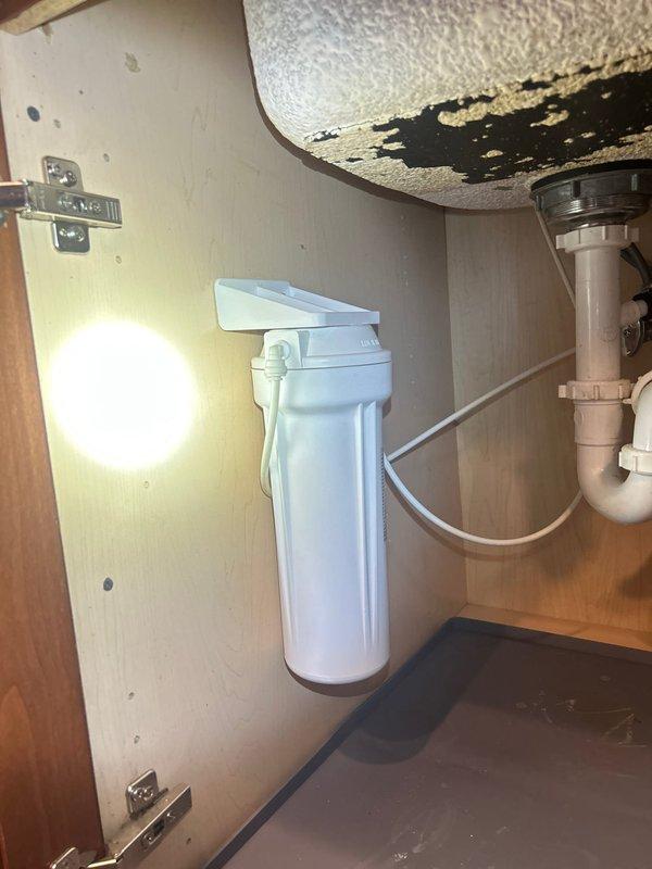 Installed under-sink water filtration system with white cylindrical filter housing and connected supply lines to existing plumbing beneath kitchen sink. System was mounted vertically to cabinet wall with proper tubing connections integrated into the cold water supply line, ensuring filtered water delivery to the faucet while maintaining existing P-trap drainage configuration.