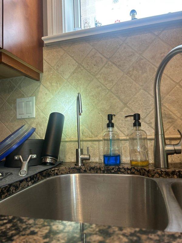 Installed under-sink water filtration system with white cylindrical filter housing and connected supply lines to existing plumbing beneath kitchen sink. System was mounted vertically to cabinet wall with proper tubing connections integrated into the cold water supply line, ensuring filtered water delivery to the faucet while maintaining existing P-trap drainage configuration.