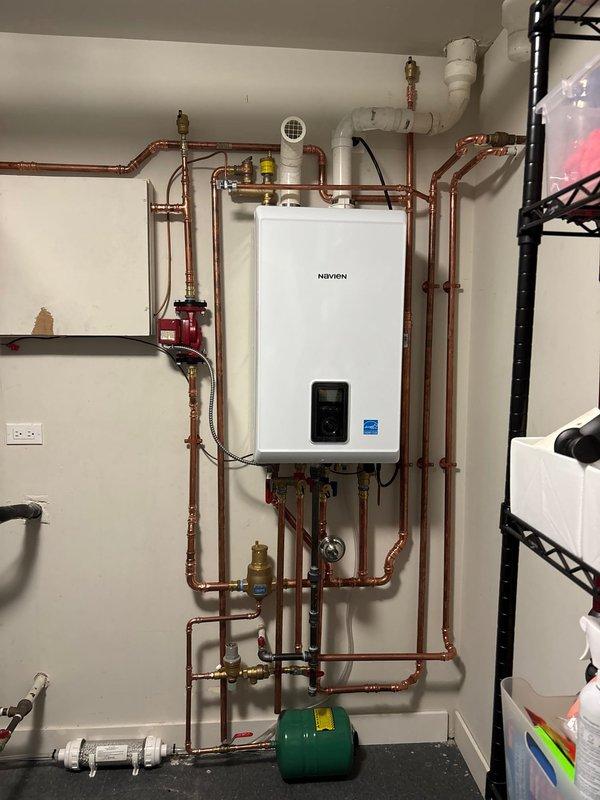 Completed professional installation of a Navien Energy Star certified tankless combi-boiler system in utility room. Installation included comprehensive copper piping network with multiple shut-off valves featuring red handles, proper venting using white PVC piping, green expansion tank mounted at floor level, and manifold distribution box with multiple zone valves. All piping runs were systematically organized with appropriate support brackets, brass fittings, and connections routed vertically and horizontally across the wall. System includes digital control panel and is fully operational with proper electrical connections established.