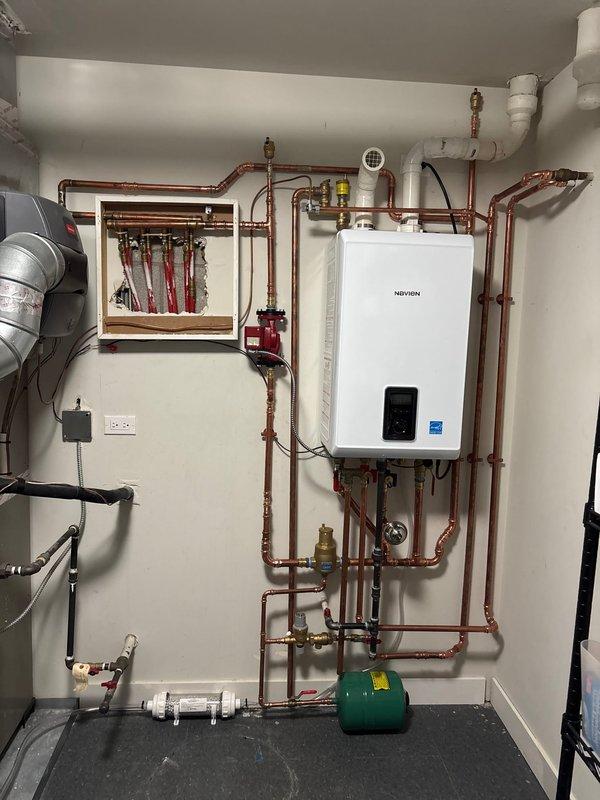 Completed professional installation of a Navien Energy Star certified tankless combi-boiler system in utility room. Installation included comprehensive copper piping network with multiple shut-off valves featuring red handles, proper venting using white PVC piping, green expansion tank mounted at floor level, and manifold distribution box with multiple zone valves. All piping runs were systematically organized with appropriate support brackets, brass fittings, and connections routed vertically and horizontally across the wall. System includes digital control panel and is fully operational with proper electrical connections established.
