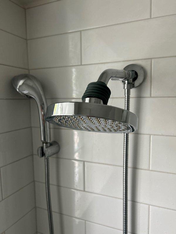 Installed customer-supplied bathroom shower fixtures on white subway-tiled wall. Mounted chrome shower head with rain-style spray pattern on wall arm with black rubber coupling boot, installed handheld shower wand holder with flexible braided stainless steel hose, and secured chrome tub spout with escutcheon plate and drain chain assembly. All fixtures properly sealed and tested for proper operation and water flow.