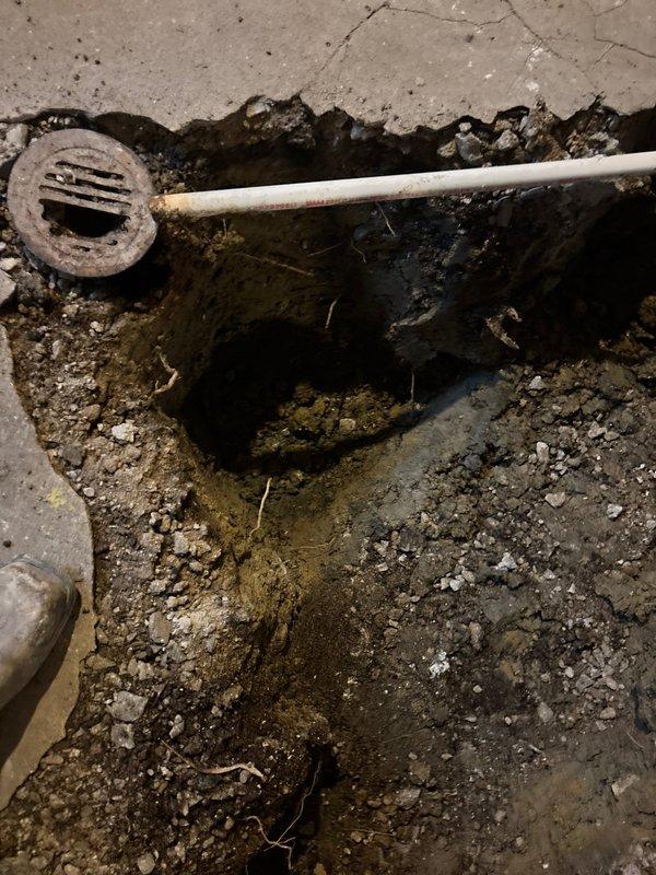 Excavated and exposed deteriorated floor drain system showing severe drainage failure with standing water accumulation and biological contamination around corroded metal drain grate. Trenched through concrete foundation to access compromised underground plumbing infrastructure, revealing saturated soil conditions and inadequate drainage beneath slab. Documented existing conditions including failed PVC drain lines, standing water in excavated cavity, and extensive moisture damage to surrounding concrete surfaces requiring comprehensive drain system replacement and proper grading restoration.