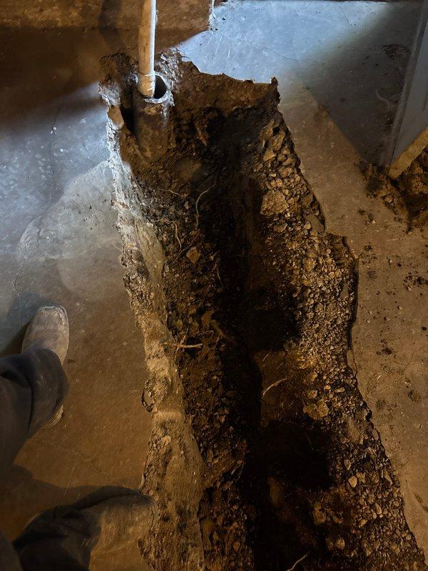 Excavated and exposed deteriorated floor drain system showing severe drainage failure with standing water accumulation and biological contamination around corroded metal drain grate. Trenched through concrete foundation to access compromised underground plumbing infrastructure, revealing saturated soil conditions and inadequate drainage beneath slab. Documented existing conditions including failed PVC drain lines, standing water in excavated cavity, and extensive moisture damage to surrounding concrete surfaces requiring comprehensive drain system replacement and proper grading restoration.