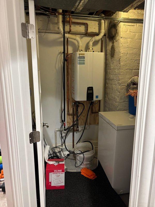 Performed camera inspection of tankless water heater installation and associated plumbing connections in utility closet. Documented wall-mounted Energy Star certified unit with copper supply lines, white PEX piping, black flexible gas lines, and multiple shut-off valves including red-handled isolation valve. Photographed complete system configuration mounted on plywood backing, capturing all pipe routing, fittings, and connections for service records.