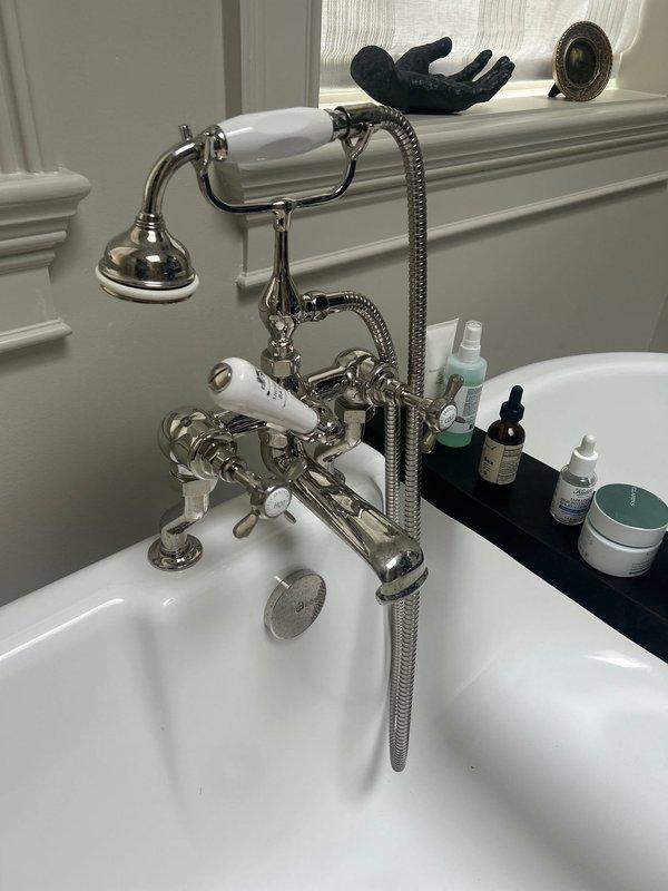 Installed handheld shower attachment with chrome mounting bracket and flexible braided metal hose on white tiled shower wall. Serviced vintage-style deck-mounted bathtub faucet assembly with polished chrome fixtures, white porcelain handle accents, and handheld shower component on freestanding tub, ensuring all connections were secure and fixtures were operating properly.
