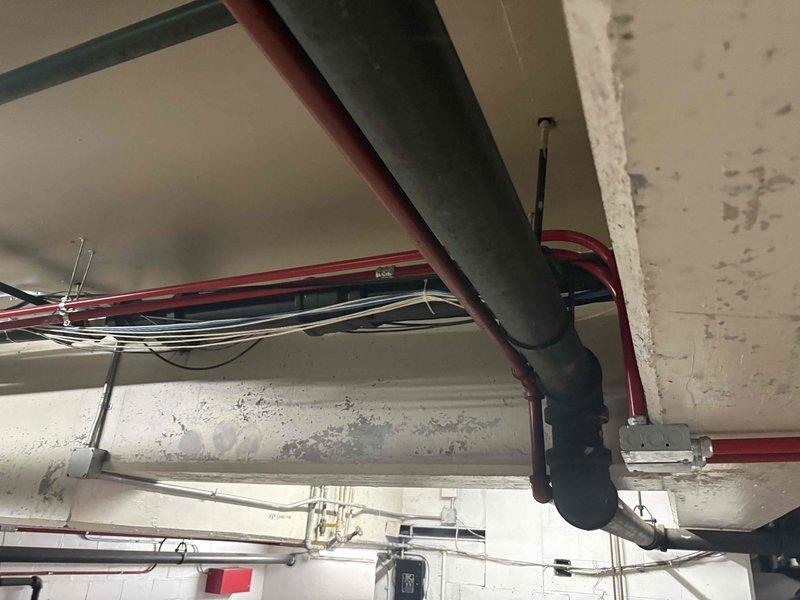 Conducted comprehensive walk-through of commercial building mechanical space to assess existing piping infrastructure and develop material list for upcoming installation. Documented overhead ceiling-mounted systems including large-diameter ductwork, insulated pipes, red-painted conduit lines, electrical wiring bundles, and associated mounting hardware and brackets in parking garage area to determine scope of work and required materials for piping installation project.