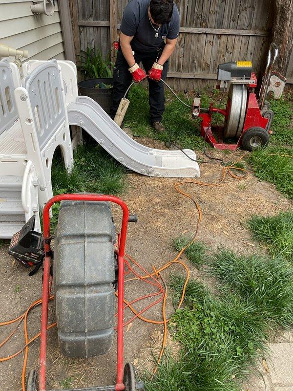 Performed sewer line rodding and camera inspection for residential property in preparation for home sale. Technician utilized motorized drain cleaning equipment with cable drum to clear the main sewer line, accessing the system through an exterior cleanout in the backyard. Following the mechanical cleaning, a comprehensive video camera inspection was conducted to document the interior condition of the sewer line and verify proper flow. Equipment setup included positioning the power rodding machine on the concrete patio area with appropriate safety protocols. Interior basement plumbing connections and floor drain access points were inspected as part of the comprehensive evaluation. Documentation completed through photographic evidence of equipment deployment and work areas to support the real estate transaction requirements.