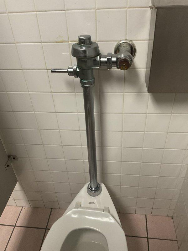 Responded to priority service call for leaking commercial toilet flush valve in women's restroom first stall. Upon inspection, identified leak originating from wall-mounted flush valve assembly on aging institutional-style toilet fixture in fifty-year-old building. Assessed flush valve mechanism, supply line connections, and associated components. Documented existing conditions showing worn chrome and gunmetal-colored flush valve body with manual flush handle, exposed piping, and signs of age-related deterioration. Determined leak source and performed necessary repairs to restore proper function and eliminate water loss.