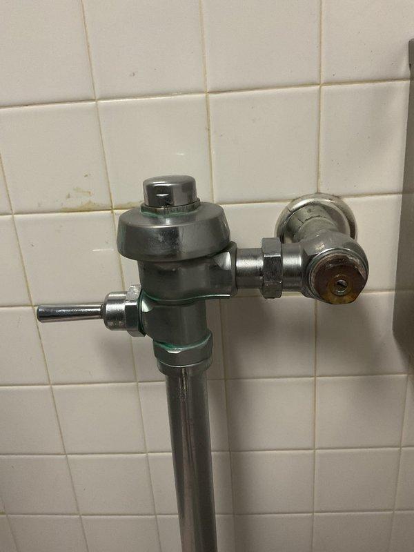 Responded to priority service call for leaking commercial toilet flush valve in women's restroom first stall. Upon inspection, identified leak originating from wall-mounted flush valve assembly on aging institutional-style toilet fixture in fifty-year-old building. Assessed flush valve mechanism, supply line connections, and associated components. Documented existing conditions showing worn chrome and gunmetal-colored flush valve body with manual flush handle, exposed piping, and signs of age-related deterioration. Determined leak source and performed necessary repairs to restore proper function and eliminate water loss.