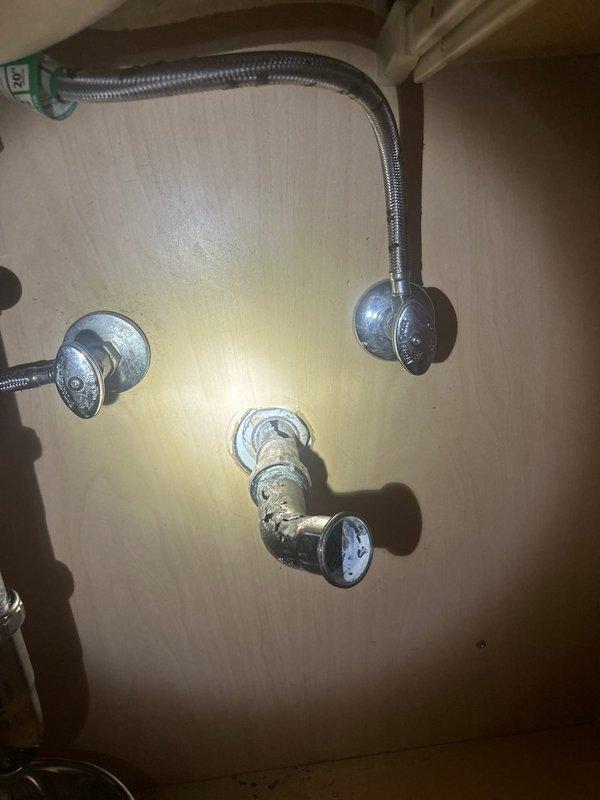 Responded to emergency call for burst pipe leaking under bathroom sink. Upon inspection, identified compromised main drainage pipe in bathroom area and examined P-trap assemblies on both vanity sink and kitchen sink as requested. Assessed plumbing fixtures including flexible braided supply lines, chrome P-trap connections, and drain assemblies for signs of failure or deterioration. Documented existing conditions showing water staining on wall surfaces near plumbing connections and verified integrity of white plastic drain components with chrome fittings. Positioned collection basin beneath affected area to prevent further water damage during assessment.
