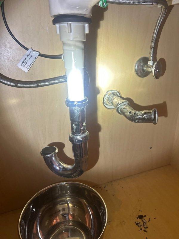 Responded to emergency call for burst pipe leaking under bathroom sink. Upon inspection, identified compromised main drainage pipe in bathroom area and examined P-trap assemblies on both vanity sink and kitchen sink as requested. Assessed plumbing fixtures including flexible braided supply lines, chrome P-trap connections, and drain assemblies for signs of failure or deterioration. Documented existing conditions showing water staining on wall surfaces near plumbing connections and verified integrity of white plastic drain components with chrome fittings. Positioned collection basin beneath affected area to prevent further water damage during assessment.