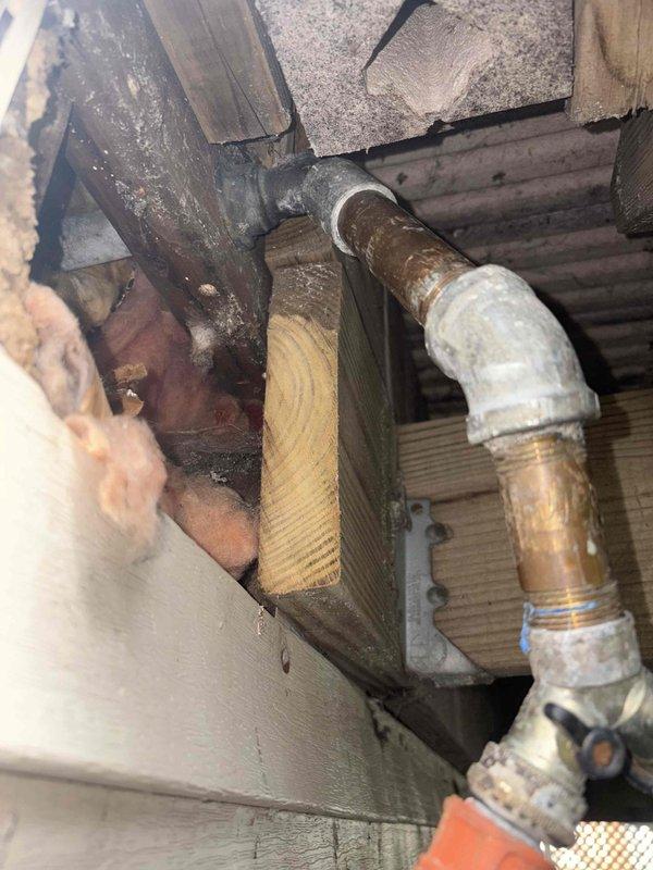 Responded to a priority call for a non-functional exterior plumbing connection that had been inoperative for approximately six months at a century-old residence. Upon inspection, accessed the crawl space and vertical utility chase to assess the damaged piping system. Found severely corroded copper piping with extensive oxidation and greenish-brown patina throughout the affected section, indicating prolonged exposure and deterioration. The compromised pipe showed threaded connections with visible corrosion at junction points and a white elbow fitting. Evaluated the routing of the existing plumbing line through the structural framing members and documented the condition of the installation for repair planning. Photographed the corroded copper pipe, deteriorated fittings, and surrounding structural components including wooden framing, insulation, and the vertical service corridor to provide a complete assessment of the repair scope required to restore functionality to the exterior water c