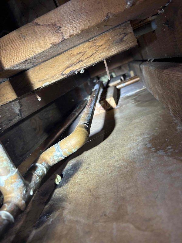 Responded to water leak originating from beneath concrete slab and manifesting in basement area of fifty-year-old residential property. Upon investigation, accessed wall cavity and exposed deteriorated copper plumbing system showing extensive corrosion with characteristic green and blue-green patina indicative of oxidized copper and prolonged moisture exposure. Identified compromised pipe sections with visible rust-colored discoloration and oxidation at horizontal runs and elbow joints within bathroom wall assembly.

Inspected subfloor structural area and located additional corroded copper piping running through floor joists with deteriorated insulation wrapping. Documented condition of aged plumbing infrastructure showing signs of long-term water intrusion and material degradation. Photographed damaged drywall sections, exposed pipe connections, and compromised wooden structural members exhibiting moisture staining and accumulated debris. Assessment confirmed active leak from corroded