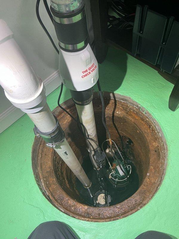 Responded to afternoon appointment for catch basin cover assessment and service. Upon arrival, accessed utility room where sump pump basin was located in floor with bright green painted surface. Inspected existing catch basin installation featuring rusted metal rim and dual white PVC pipe configuration with quiet check valve system. Basin contained operational sump pump mechanism with electrical connections and showed signs of age-related corrosion on metal components. Documented current condition of installation including water level, pipe positioning, and overall basin integrity for maintenance records.