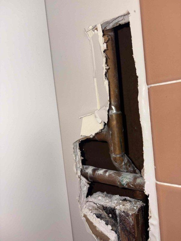 Responded to emergency plumbing repair involving severely corroded copper supply lines concealed within bathroom wall. Upon investigation, discovered extensive deterioration of copper piping with visible oxidation, greenish-blue patina, and white mineral deposits indicating chronic moisture exposure and potential leak activity. Removed damaged section of drywall to access plumbing system and assess full extent of corrosion at pipe joints and fittings. The aged copper pipes displayed significant tarnishing and compromised integrity requiring immediate attention to prevent catastrophic failure and water damage. Documented existing conditions with photographic evidence showing corroded pipe connections, mineral buildup, and deteriorated plumbing components within wall cavity adjacent to tiled bathroom surfaces.