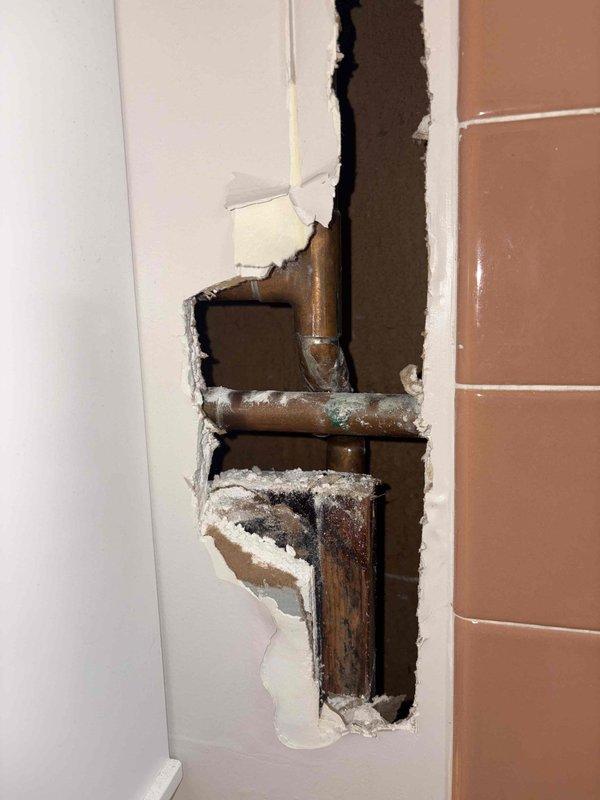 Responded to emergency plumbing repair involving severely corroded copper supply lines concealed within bathroom wall. Upon investigation, discovered extensive deterioration of copper piping with visible oxidation, greenish-blue patina, and white mineral deposits indicating chronic moisture exposure and potential leak activity. Removed damaged section of drywall to access plumbing system and assess full extent of corrosion at pipe joints and fittings. The aged copper pipes displayed significant tarnishing and compromised integrity requiring immediate attention to prevent catastrophic failure and water damage. Documented existing conditions with photographic evidence showing corroded pipe connections, mineral buildup, and deteriorated plumbing components within wall cavity adjacent to tiled bathroom surfaces.
