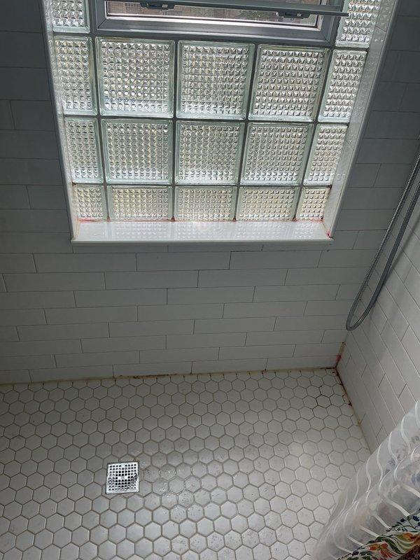 Conducted warranty inspection of bathroom shower area following customer report of tiles detaching from surfaces. Upon examination, found white hexagonal floor tiles and subway wall tiles intact and properly adhered. However, identified significant rust-colored staining and discoloration around glass block window frame corners and floor-to-wall junctions, indicating potential moisture infiltration issues. The discoloration patterns suggest water may be penetrating behind the tile work or through window frame seals, which could compromise tile adhesion over time if left unaddressed. Documented conditions with photographs for warranty evaluation purposes.