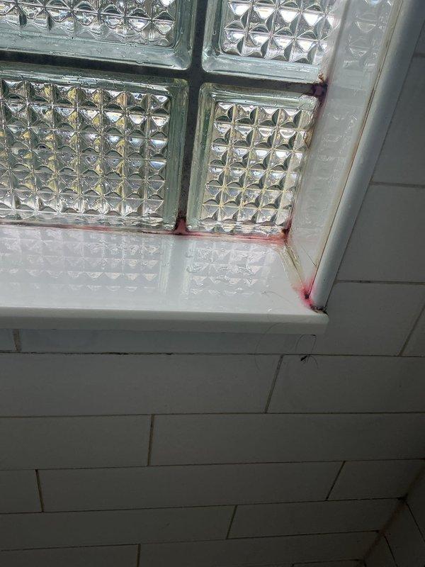 Conducted warranty inspection of bathroom shower area following customer report of tiles detaching from surfaces. Upon examination, found white hexagonal floor tiles and subway wall tiles intact and properly adhered. However, identified significant rust-colored staining and discoloration around glass block window frame corners and floor-to-wall junctions, indicating potential moisture infiltration issues. The discoloration patterns suggest water may be penetrating behind the tile work or through window frame seals, which could compromise tile adhesion over time if left unaddressed. Documented conditions with photographs for warranty evaluation purposes.
