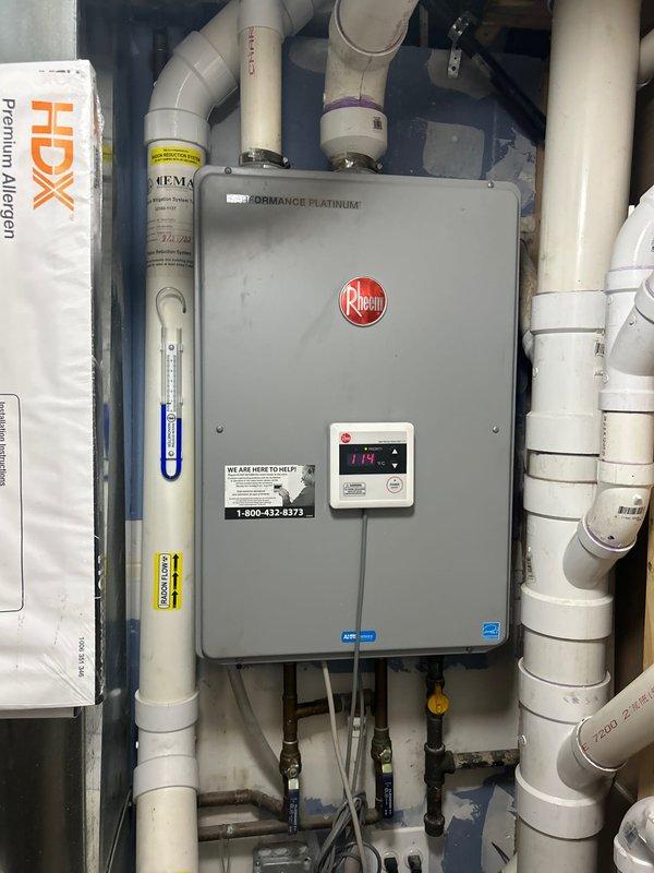 Completed installation and inspection of Rheem Performance Platinum tankless water heater system with associated plumbing connections. Verified proper operation of water supply shut-off valves beneath sink cabinet, documented existing water staining on cabinet interior surfaces, and confirmed temperature settings at 119°F on digital display. All PVC piping connections, inlet/outlet lines, and safety components including pressure relief valve and temperature gauge were inspected and photographed for service records.