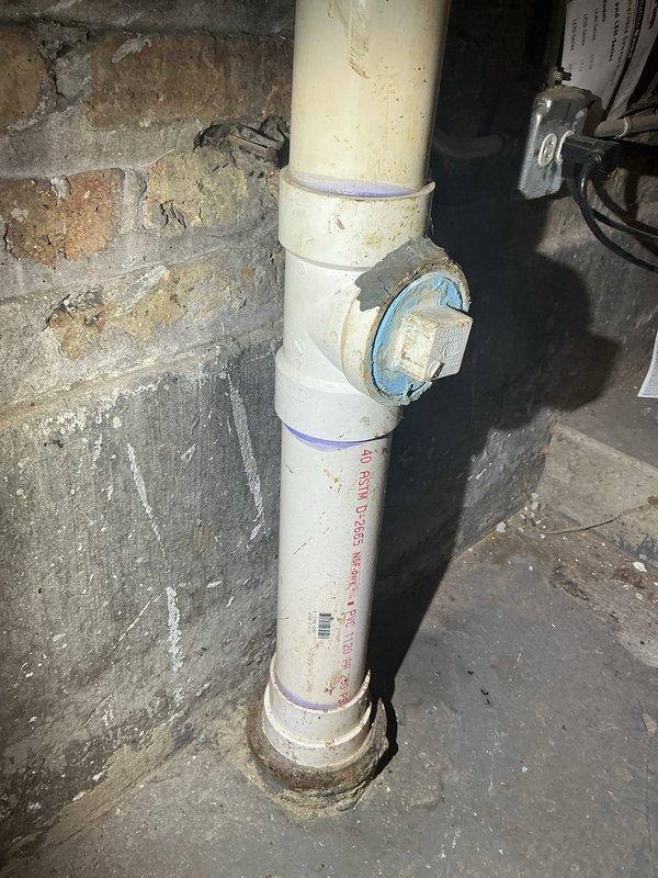 Completed callback to assess cleanout condition and verify scope of previous maintenance service. Upon inspection, documented damaged cleanout fitting on vertical PVC drain line in basement utility area. Photographed the blue-capped cleanout access port showing visible wear and the surrounding plumbing infrastructure including vertical white PVC drainage pipe with manufacturer markings and purple specification bands. Contacted service coordinator to confirm whether hydrojetting was included in prior maintenance visit, as this information is necessary to determine if cleanout damage occurred during previous service or was pre-existing condition.