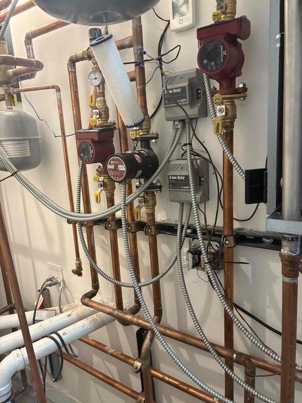 Responded to service call for loss of hot water and multiple plumbing concerns. Inspected existing tanked water heater installed in 2017 and confirmed ignition failure on unit. Attempted reignition procedures without success, determining unit required replacement. Water heater serves both domestic hot water and ice melt system through hydronic heating zones controlled by zone relays and multiple circulator pumps. 

Assessed copper supply lines, zone control valves, and circulator pump operation throughout the multi-zone heating distribution system. Photographed existing installation including floor penetrations with copper refrigerant lines, HVAC venting, and spray foam air sealing. Documented complete hydronic system configuration showing red circulator pumps, gray zone relay control boxes, pressure gauge, expansion tank, brass valves and fittings, and PVC drain lines.

Evaluated reported pipe burst at exterior spigot requiring repair. Provided estimate for sump pump installation with