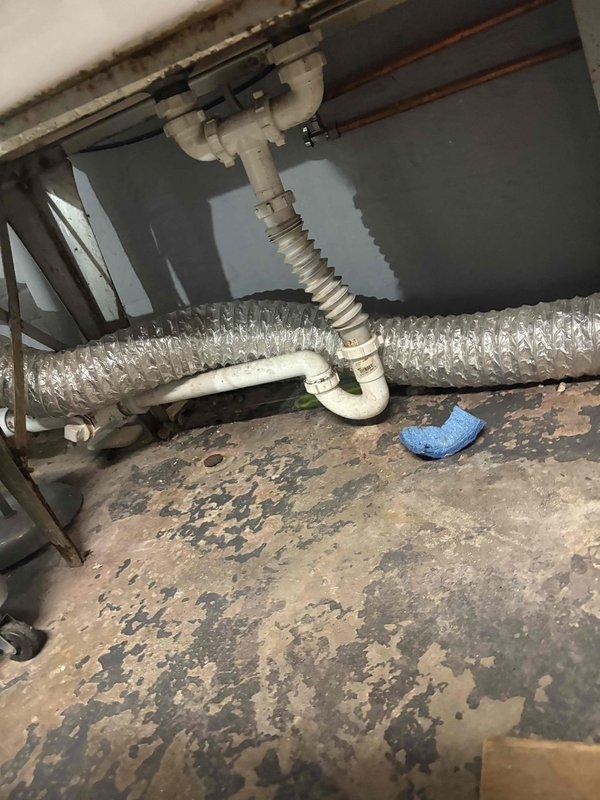 Responded to water leak originating from main plumbing stack. Upon inspection, discovered active leak causing extensive moisture damage to subflooring beneath sink cabinet and concrete basement floor below. The deteriorated P-trap assembly and corrugated drain hose connections showed signs of failure, contributing to water intrusion. Documented existing damage including severely compromised plywood subflooring with advanced peeling and discoloration, as well as substantial water staining on basement concrete floor surrounding the vertical drain stack penetration. Photographed affected areas showing damaged plumbing components, deteriorated flooring materials, and moisture patterns for repair assessment and future remediation planning.