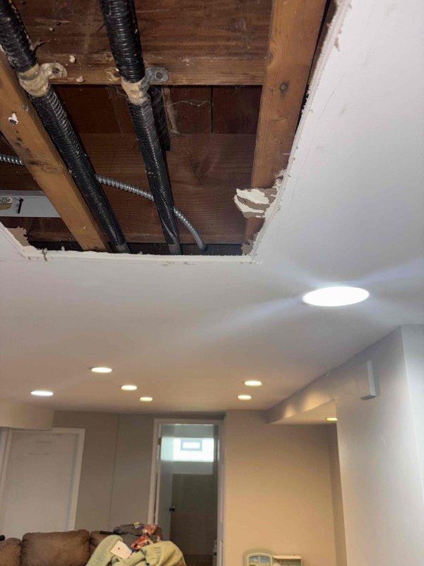 Responded to emergency plumbing service call for burst pipe repair in residential basement living area. Created ceiling and wall access points to reach compromised drain system components. Replaced severely corroded metal drain trap assembly showing significant oxidation and mineral buildup with new PVC P-trap configuration. Applied proper primer and secured all connections to code. Addressed water damage concerns by exposing affected structural areas including ceiling joists and wall cavities to ensure proper assessment and drying. Rerouted and secured existing flexible conduits during repair process. Verified all drain connections for proper flow and seal integrity upon completion.