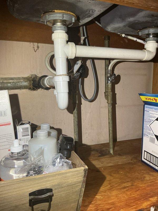 Completed under-sink plumbing inspection and repair service for double-basin kitchen sink installation. Serviced P-trap drainage system with white PVC piping connecting both sink drains, including horizontal cross-connection and vertical drain assemblies. Addressed corroded copper supply line pipes showing significant patina and discoloration, with new flexible braided supply lines installed and connected to chrome shut-off valves. Verified proper connections for garbage disposal and dishwasher drain lines with secure metal clamps.