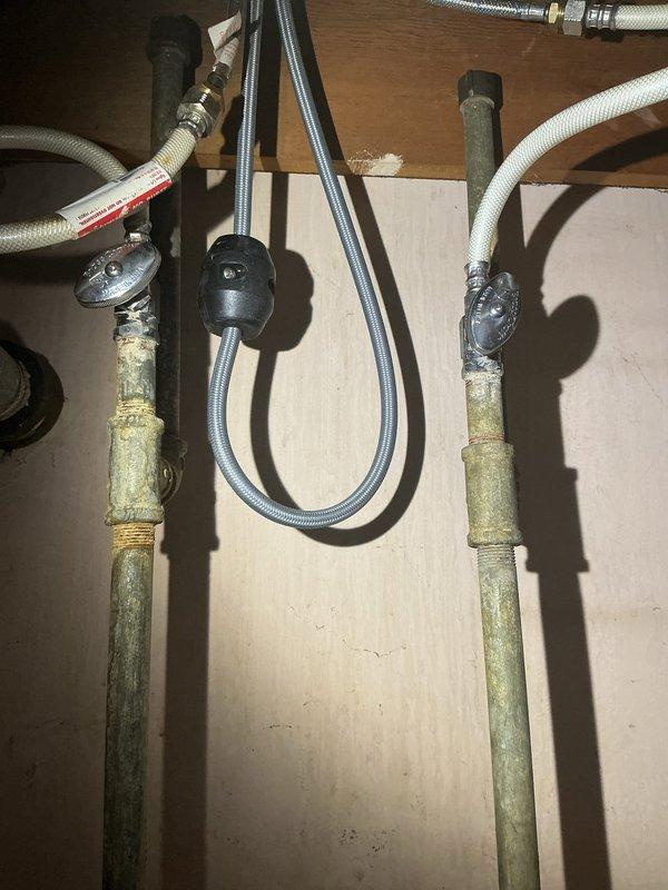 Completed under-sink plumbing inspection and repair service for double-basin kitchen sink installation. Serviced P-trap drainage system with white PVC piping connecting both sink drains, including horizontal cross-connection and vertical drain assemblies. Addressed corroded copper supply line pipes showing significant patina and discoloration, with new flexible braided supply lines installed and connected to chrome shut-off valves. Verified proper connections for garbage disposal and dishwasher drain lines with secure metal clamps.
