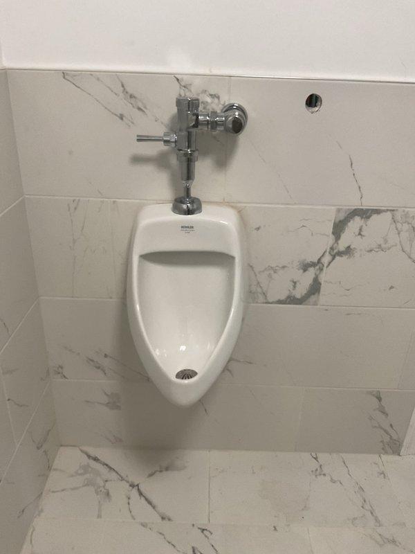 Completed finish plumbing installation in commercial restroom, including wall-mounted urinal with chrome flush valve and handle assembly. Installation followed rough plumbing work previously performed, with all fixtures mounted to marble-patterned tile surfaces and connected to existing PVC and copper supply lines.