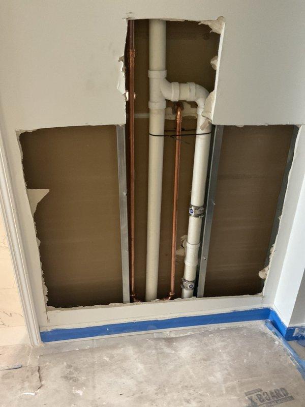 Completed finish plumbing installation in commercial restroom, including wall-mounted urinal with chrome flush valve and handle assembly. Installation followed rough plumbing work previously performed, with all fixtures mounted to marble-patterned tile surfaces and connected to existing PVC and copper supply lines.
