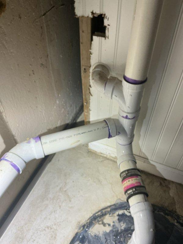 Responded to morning appointment request for plumbing repair consultation in basement utility area. Upon inspection, identified significant wall damage in corner space where white PVC drainage piping is installed, with irregular opening exposing underlying materials and brown discoloration around damaged section. Documented existing plumbing configuration including multiple elbow joints, T-joint connections, and vertical/horizontal pipe runs with purple primer markings and color-coded identification bands. Photographed complete basement utility room setup showing exposed ceiling joists, concrete block walls, white PVC drainage and ventilation systems, cylindrical black water treatment device mounted on vertical pipe, utility sink beneath window, and patterned floor covering with visible staining. Completed visual assessment of plumbing assembly condition and wall integrity for repair recommendations.