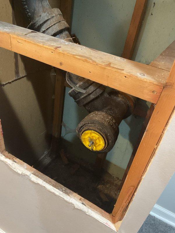 Responded to warranty callback for toilet backup into bathtub occurring two hours after completion of major plumbing work. Accessed cast iron drain and vent system through wall cavity to investigate drainage obstruction. Inspected deteriorated piping system showing significant corrosion and age-related wear, with visible rust accumulation on vertical and horizontal drain lines at pipe junction with yellow-capped valve assembly.