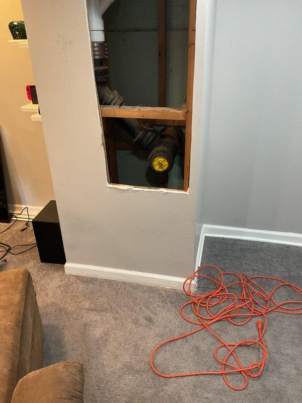 Responded to warranty callback for toilet backup into bathtub occurring two hours after completion of major plumbing work. Accessed cast iron drain and vent system through wall cavity to investigate drainage obstruction. Inspected deteriorated piping system showing significant corrosion and age-related wear, with visible rust accumulation on vertical and horizontal drain lines at pipe junction with yellow-capped valve assembly.