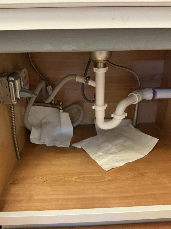 Responded to service call for kitchen faucet leak and under-sink leak in powder room. Inspected kitchen sink cabinet plumbing system, identifying moisture issues in P-trap assembly and supply line connections. Placed absorbent materials beneath plumbing fixtures to monitor leak sources and protect cabinet interior. Documented existing plumbing configuration including white PVC P-trap, metallic drain basket, chrome flexible supply lines, and associated dishwasher/disposal connections for further diagnostic assessment and repair recommendations.