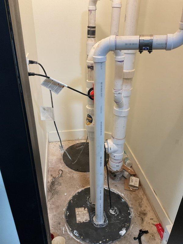 Completed installation of new sump pump system with dual basin configuration and PVC discharge piping. The system includes properly mounted pump unit with electrical connections, vertical discharge pipe assembly with appropriate fittings and elbows, and floor-mounted basins secured in concrete for optimal drainage performance.