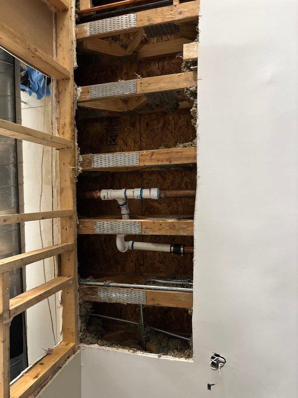Accessed interior wall cavity through drywall opening to expose horizontal framing members and plumbing infrastructure. Inspected white PVC drainage and vent system including horizontal pipe runs, T-joints, and fittings mounted between wooden joists with metal reinforcement plates. Performed necessary repairs to plumbing system and documented existing conditions of wall cavity, framing, and pipe configuration for service records.