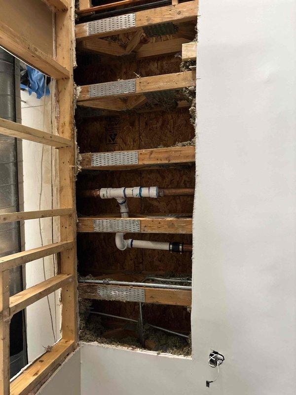 Accessed interior wall cavity through drywall opening to expose horizontal framing members and plumbing infrastructure. Inspected white PVC drainage and vent system including horizontal pipe runs, T-joints, and fittings mounted between wooden joists with metal reinforcement plates. Performed necessary repairs to plumbing system and documented existing conditions of wall cavity, framing, and pipe configuration for service records.