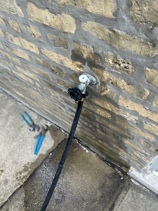 Completed installation and connection of outdoor water spigot system with chrome-finished faucet mounted to weathered wooden privacy fence. Connected hose reel assembly with functional water delivery system, ensuring proper mounting hardware and secure attachment points to accommodate exterior utility access for residential property maintenance.