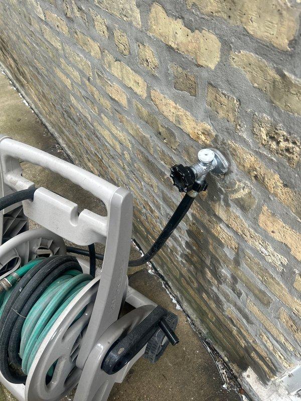 Completed installation and connection of outdoor water spigot system with chrome-finished faucet mounted to weathered wooden privacy fence. Connected hose reel assembly with functional water delivery system, ensuring proper mounting hardware and secure attachment points to accommodate exterior utility access for residential property maintenance.