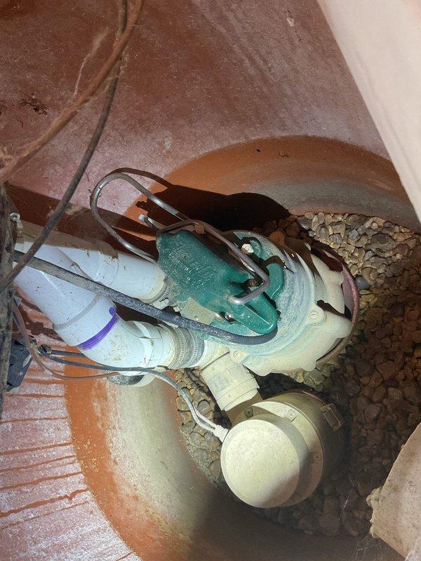 Performed comprehensive sump pump maintenance in residential basement utility area. Inspected sump pump pit, verified proper operation of green and white pump unit, and examined all electrical connections and wiring leading to the pit. Checked PVC discharge piping system, inspected pipe joints and connections including turquoise valve assembly at water heater area, and confirmed proper drainage flow. Evaluated gravel bed surrounding equipment, assessed concrete pit condition, and documented system components with photographic records to verify maintenance completion and equipment functionality.