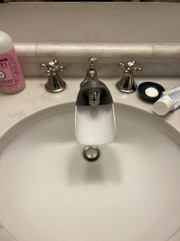 Serviced bathroom lavatory faucet with three-handle chrome fixture showing signs of age and tarnishing. Disassembled center dome cap to access valve stem assembly and inspect internal components for proper operation. Documented existing fixture condition including worn chrome finish and brass valve mechanism. Completed fixture maintenance and reassembly to restore functionality.
