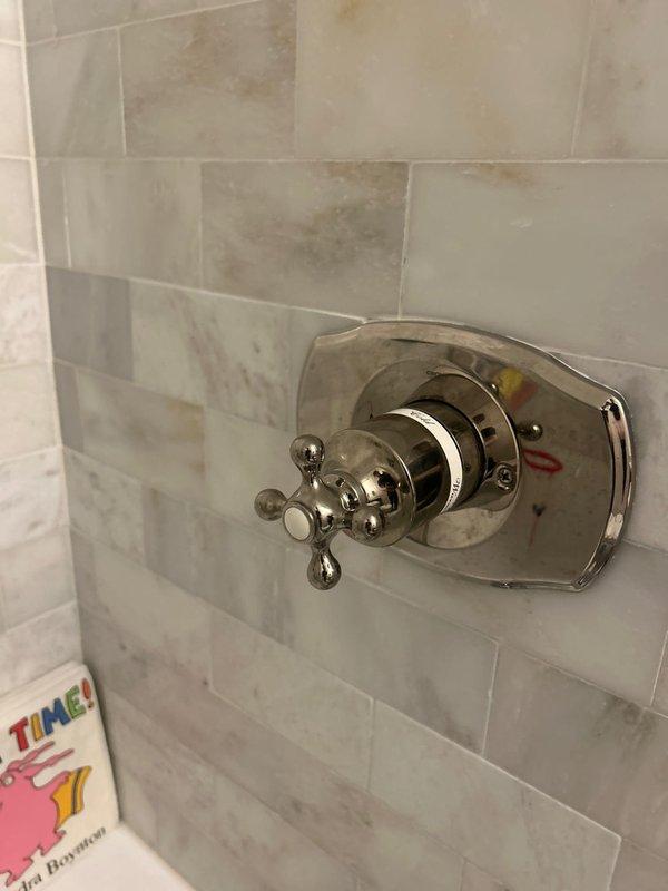 Completed installation of traditional-style chrome bathroom fixtures including a brushed nickel soap dispenser at the vanity sink and a polished chrome shower valve with cross-handle design. Both fixtures were mounted on tiled surfaces featuring subway tile pattern at sink area and decorative stone-look tile at shower location, ensuring proper alignment and secure installation for optimal functionality.