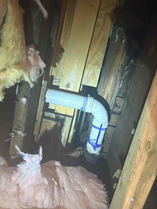 Inspected plumbing infrastructure in crawl space and identified issues with ceiling fixture mounting point showing signs of moisture intrusion and surface deterioration. Examined white PVC drainage line with visible condensation between floor joists, verified proper installation and support among structural members and fiberglass insulation. Documented existing conditions including weathered ceiling surface with crackled paint pattern around central fixture, indicating potential water damage requiring monitoring or future repair consideration.