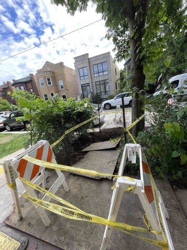 Completed excavation and repair work on residential sidewalk to access underground plumbing utilities. Excavated concrete section approximately 6-8 feet in length to expose subsurface infrastructure, installed proper safety barricades and caution tape around work zone, and provided temporary wooden walkway for pedestrian access. Work area secured with utility access covers and safety measures in place pending final restoration.