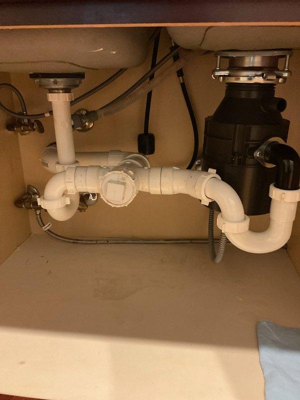 Responded to clogged kitchen sink drain and successfully cleared the blockage using rodding equipment. Accessed the drainage system through the clean-out port located in the horizontal drain line beneath the double-basin sink. The plumbing configuration consisted of white PVC piping with dual P-traps connecting to a central horizontal drain pipe, with the right basin equipped with a garbage disposal unit. Restored full drainage function to both sink basins.