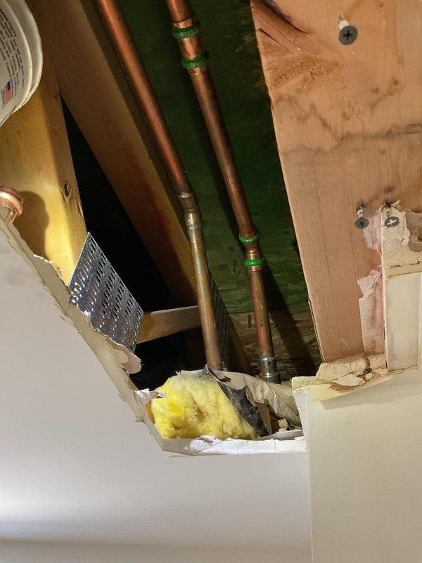 Responded to a ceiling leak originating from shower plumbing in a 20-year-old residence. Accessed ceiling cavity below bathroom to locate source of water intrusion. Inspected copper supply lines and connections, identifying compromised pipe fittings as the leak source. Created access opening in ceiling drywall to expose plumbing infrastructure and assess extent of water damage to insulation and framing. Documented existing conditions with photographs showing copper piping, mounting brackets, and saturated fiberglass insulation requiring attention.