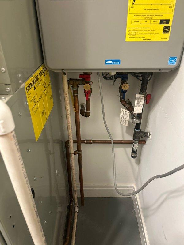 Completed installation and setup of an A.O. Smith ENERGY STAR certified tankless water heater system. Unit was properly mounted in a white metal cabinet enclosure with all necessary plumbing connections established, including copper supply lines with red and blue handled shut-off valves for hot and cold water control, white PEX flexible tubing, and white PVC drainage piping. Electrical connections were secured and all components were organized within the cabinet for optimal access and maintenance. Verified proper operation of the system and confirmed functionality with the handheld shower fixture in the adjacent bathroom, which features contemporary tile work and chrome fixtures.