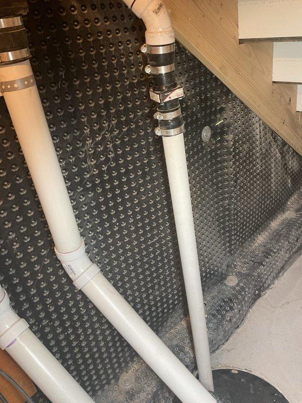 Completed site assessment for sump pump and battery backup system installation in basement crawl space. Evaluated existing plumbing infrastructure including white PVC drainage lines with proper rubber coupling connectors and metal hose clamps. Documented drainage membrane system with dimpled moisture management matting showing water accumulation patterns, indicating need for active water removal solution. Assessed electrical service availability at installation location with existing outlet box suitable for pump power connection. Photographed current conditions including floor joist clearances, pipe routing configurations, and available space for pump basin and backup battery system placement. Prepared comprehensive estimate for primary sump pump installation with battery backup system to address basement water management needs.