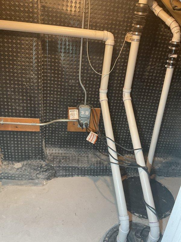 Completed site assessment for sump pump and battery backup system installation in basement crawl space. Evaluated existing plumbing infrastructure including white PVC drainage lines with proper rubber coupling connectors and metal hose clamps. Documented drainage membrane system with dimpled moisture management matting showing water accumulation patterns, indicating need for active water removal solution. Assessed electrical service availability at installation location with existing outlet box suitable for pump power connection. Photographed current conditions including floor joist clearances, pipe routing configurations, and available space for pump basin and backup battery system placement. Prepared comprehensive estimate for primary sump pump installation with battery backup system to address basement water management needs.